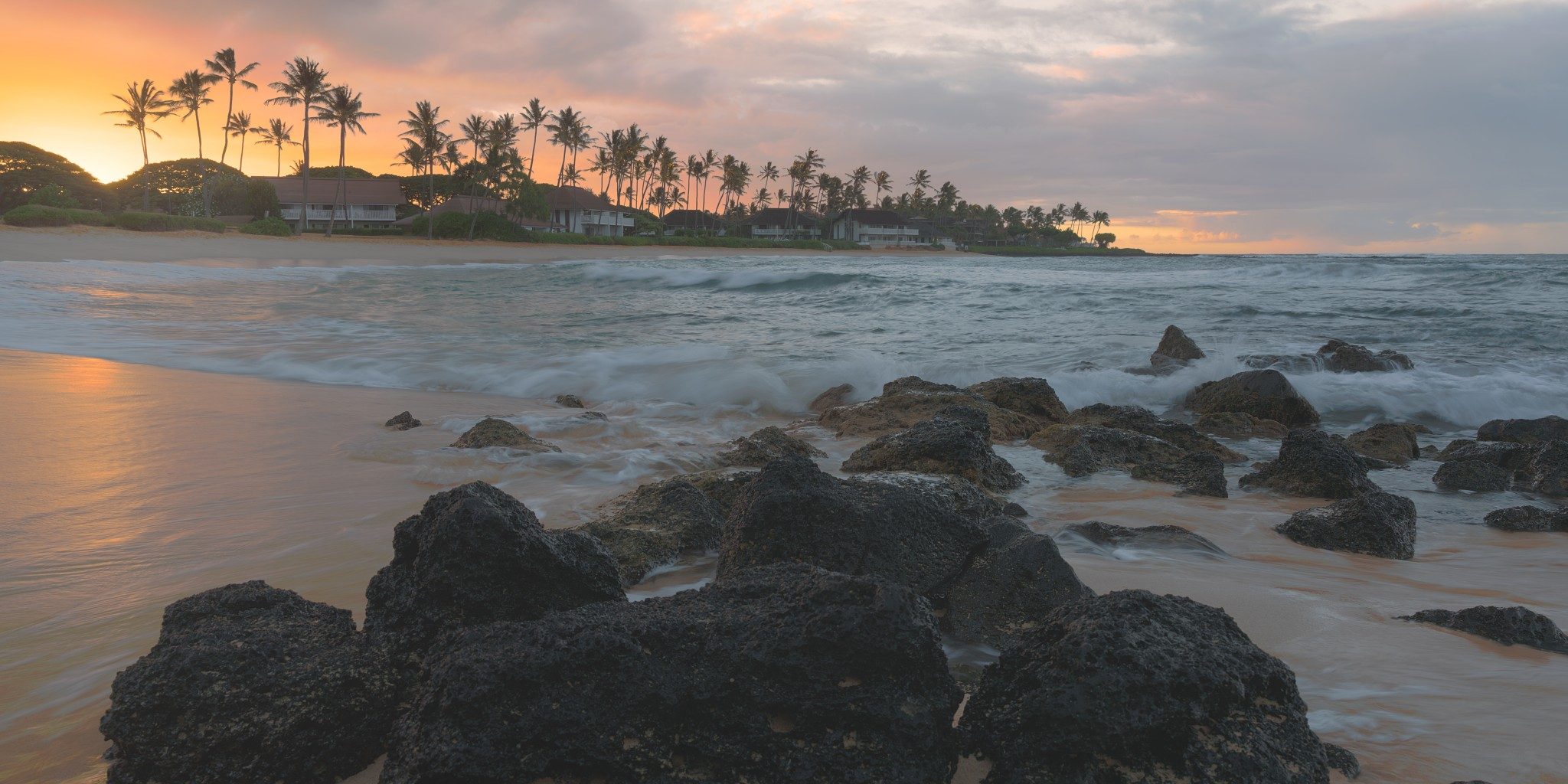 Dark lava rocks in smooth, misty water on a sandy beach at sunrise, with a line of palm trees on a point under an orange and pink sky.