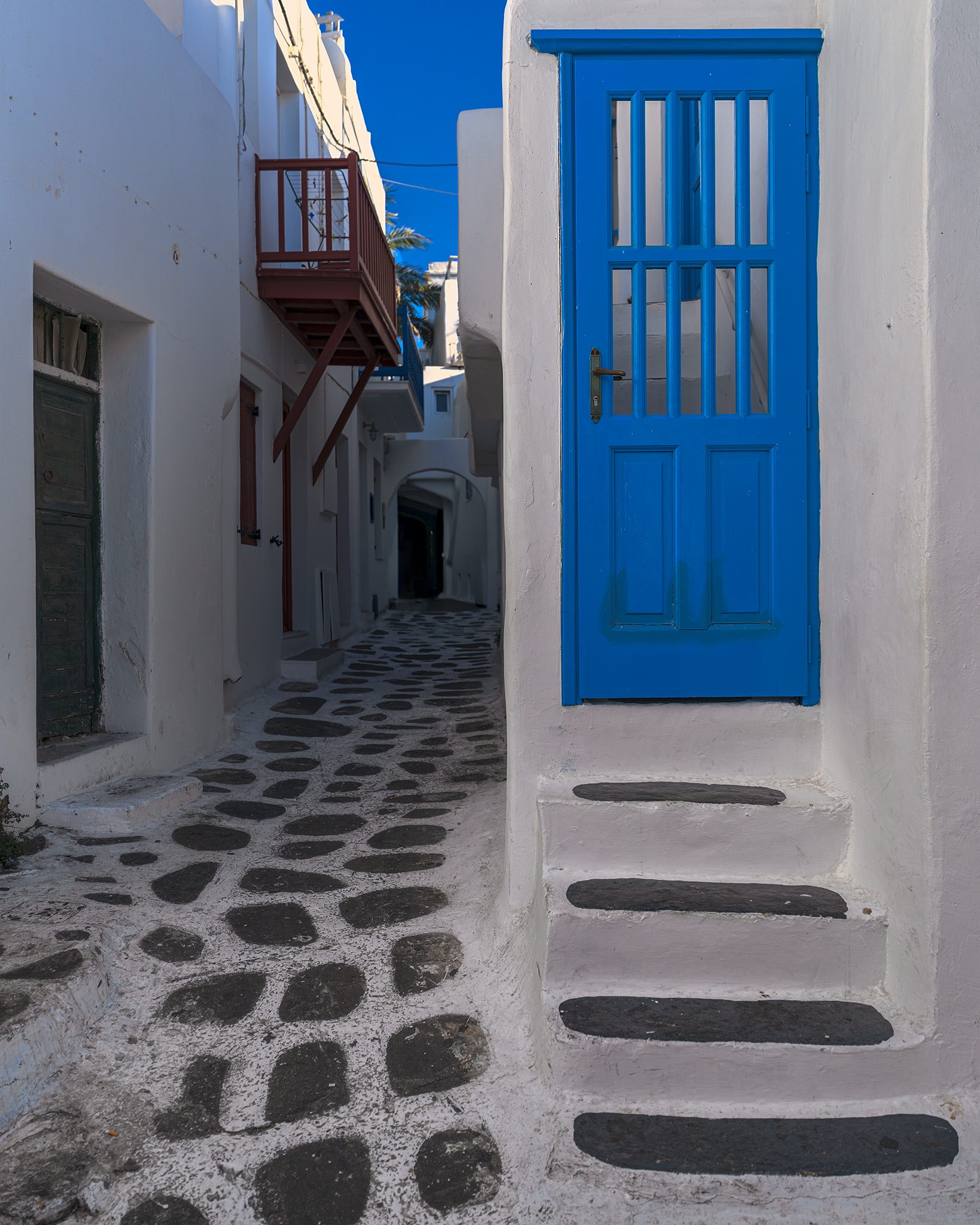 A cobalt blue paneled door at the top of whitewashed steps edged in gray, beside a narrow lane paved with white-outlined stones and a small wooden balcony.