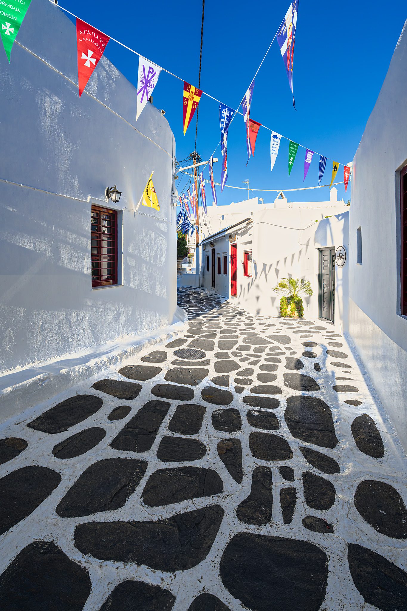 A narrow whitewashed alley with dark paving stones outlined in white, strings of colorful pennant flags crossing overhead against a deep blue sky.